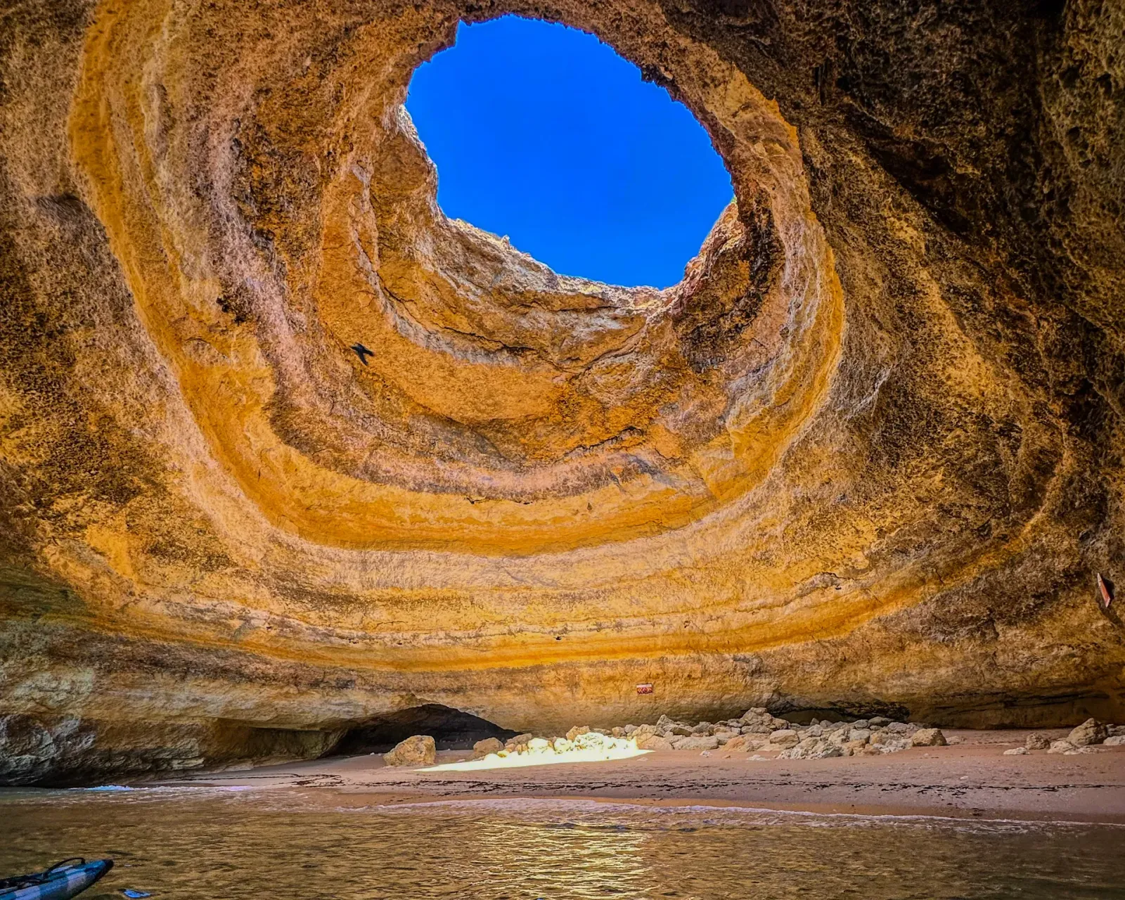Panoramic wide-angle view inside Benagil Cave showing the iconic circular skylight opening in the ceiling, golden rock walls, and turquoise water below – Algarve, Portugal