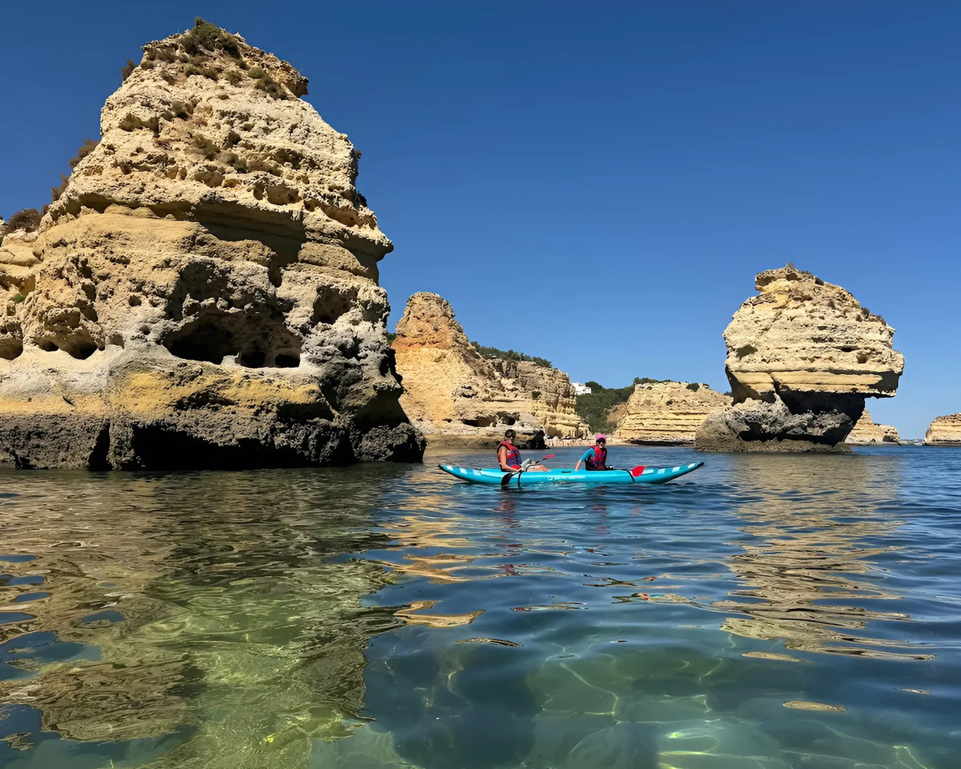 Couple in a tandem kayak navigating between tall rock pillars in crystal-clear water along the Algarve coast near Benagil