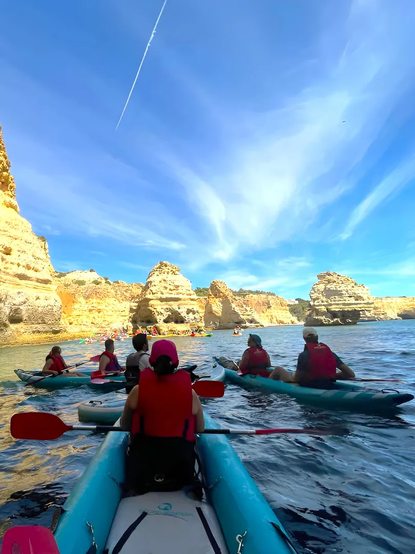 Guided kayak group paddling past dramatic Algarve rock formations with golden light reflecting off the cliff faces