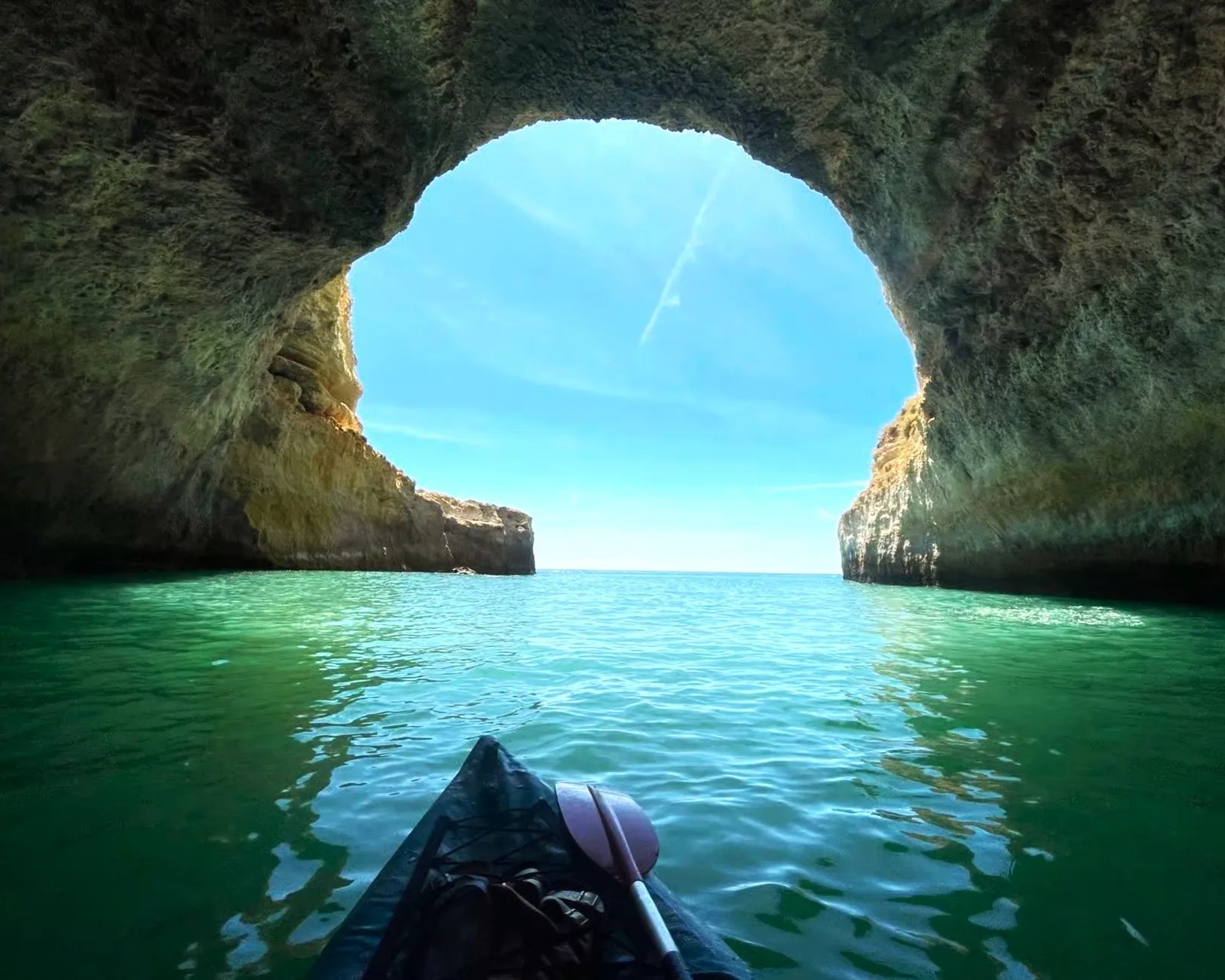 Kayak point-of-view paddling through a sea arch with vivid emerald water and rugged Algarve coastline visible through the opening